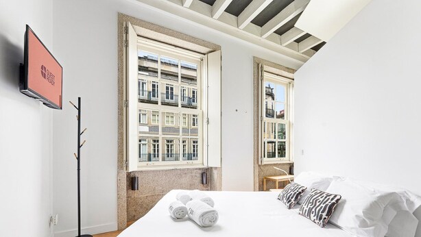 Een moderne hotelkamer met een groot bed, twee ramen met uitzicht op een gebouw, een kapstok en een flatscreen televisie aan de muur.