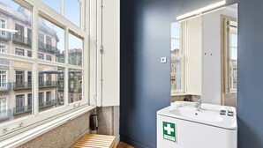 Een badkamer met een groot raam, een wastafel met een bordje voor eerste hulp en een houten bankje.
