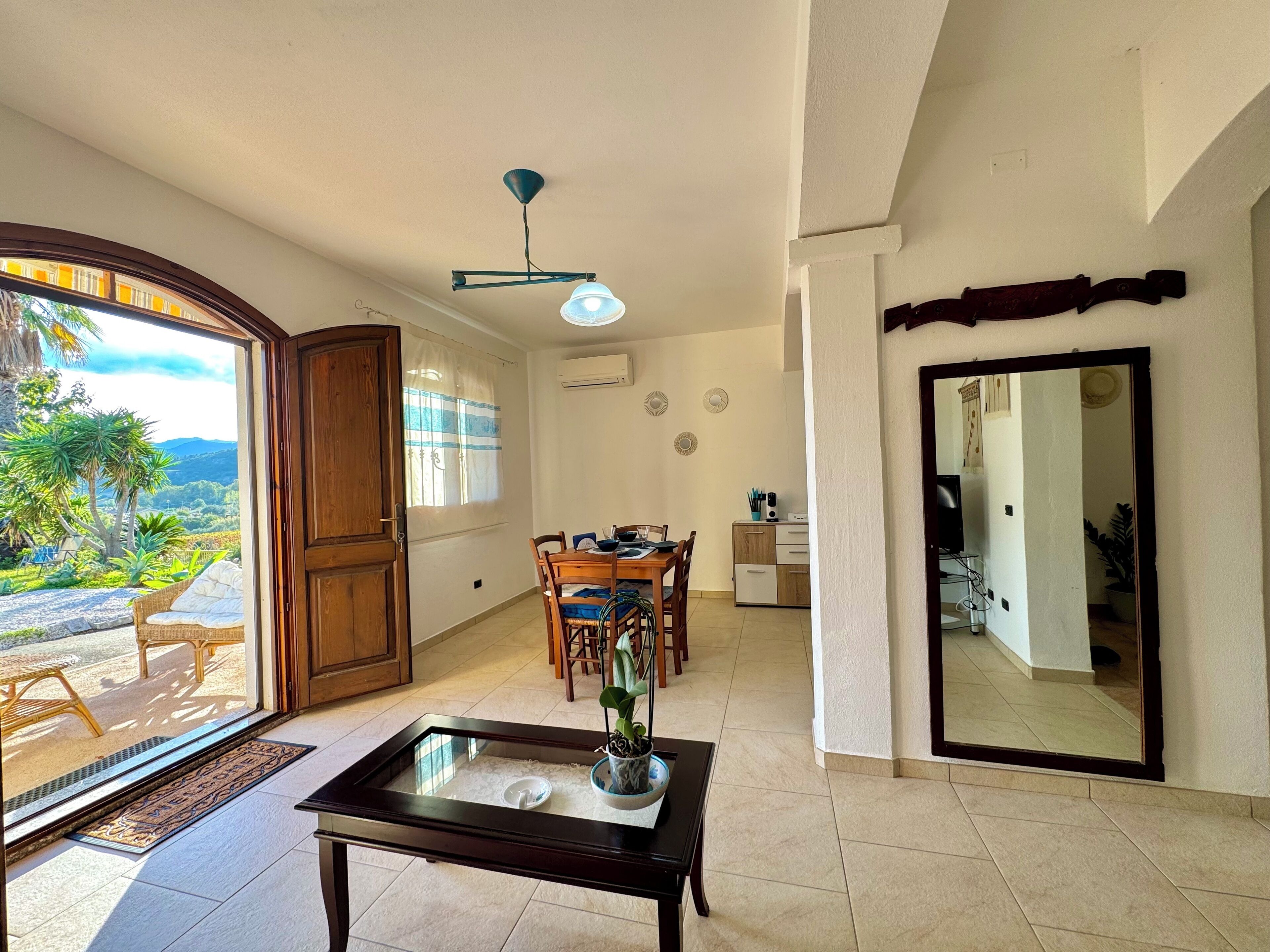 Amplia sala de estar con mesa de comedor, un espejo grande y vistas a un paisaje exuberante a través de una puerta corrediza.