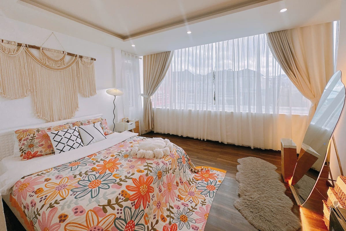 A bedroom with a floral bedspread, a large window with curtains, a bedside lamp, and a mirror.