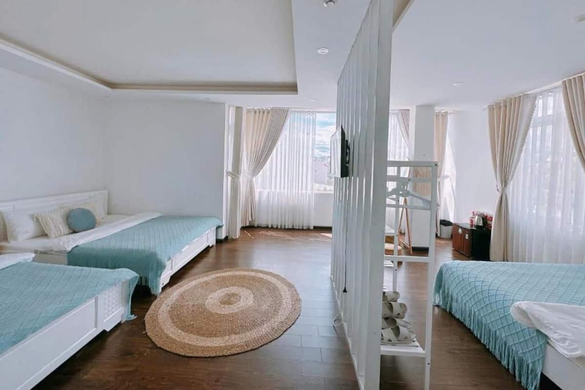 A hotel room with two single beds, a large woven rug, a white wardrobe, and a view of the city through large windows.