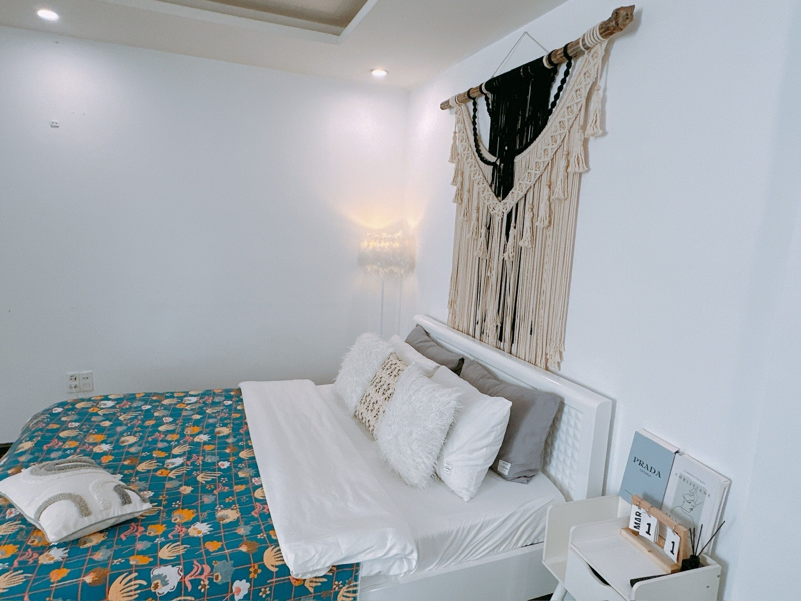A bedroom with a bed, a wall-mounted macrame piece, a bedside table with a clock, and a wall-mounted light.
