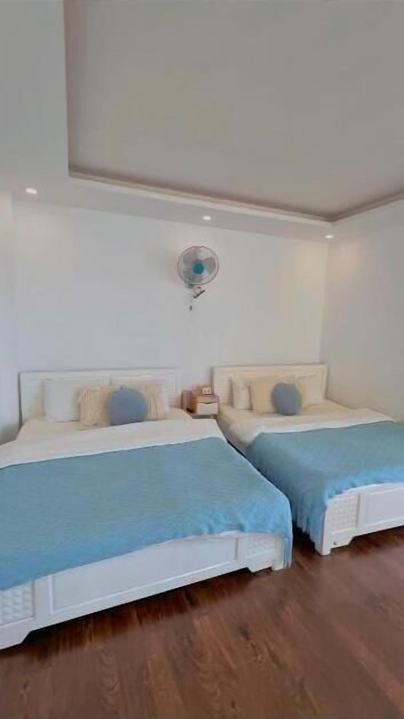 A bedroom with two beds, each with a blue bedspread and white headboards. There is a small bedside table with a lamp on each bed. The room has white walls and a wooden floor.