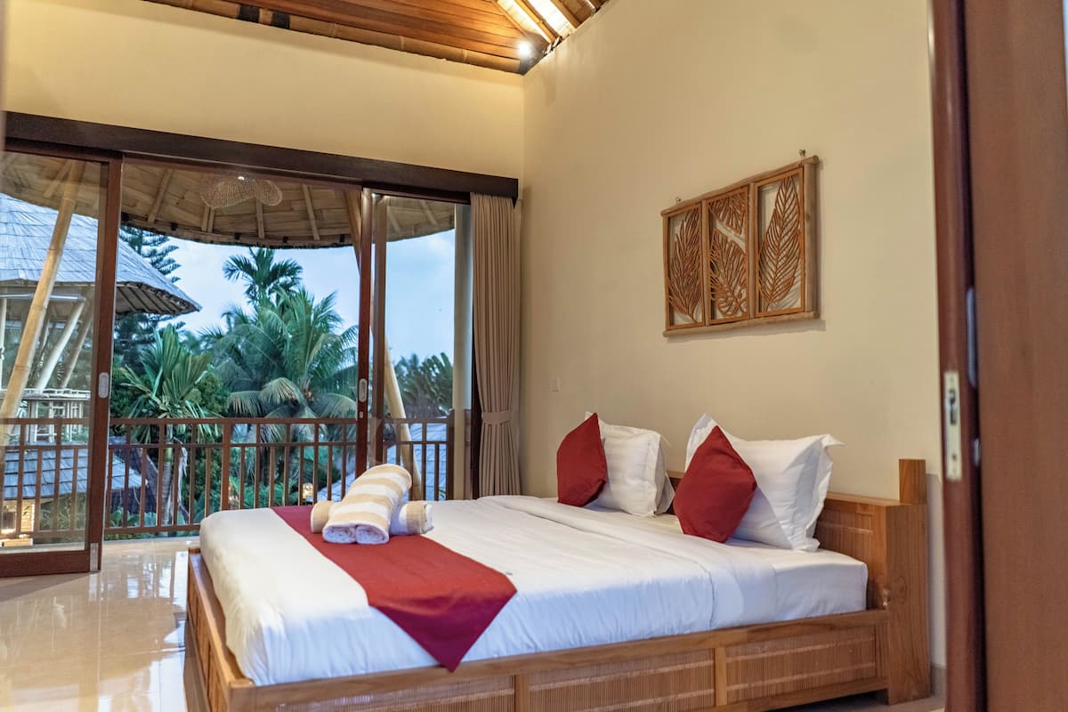 A bedroom with a large bed, a balcony with a view of palm trees, and a wooden ceiling.