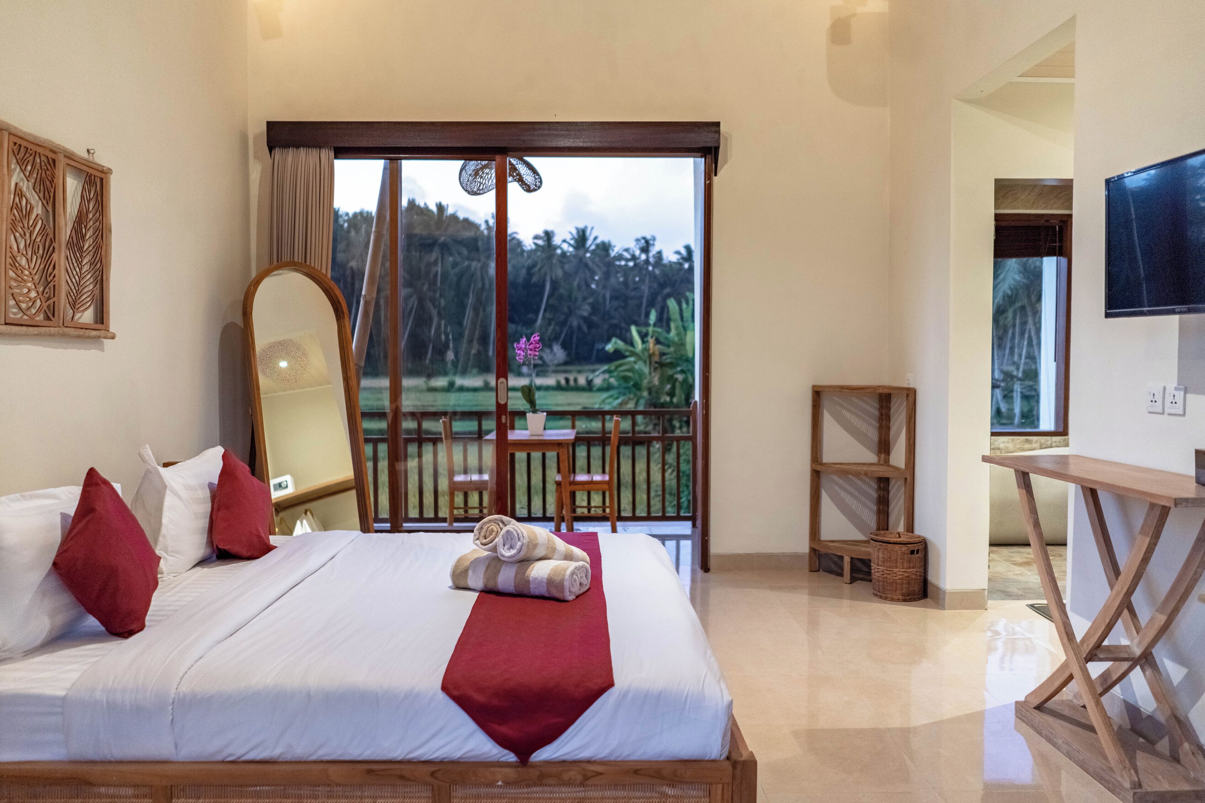 Une chambre d’hôtel avec un lit, un grand miroir, une télévision et un balcon offrant une vue sur un paysage tropical.