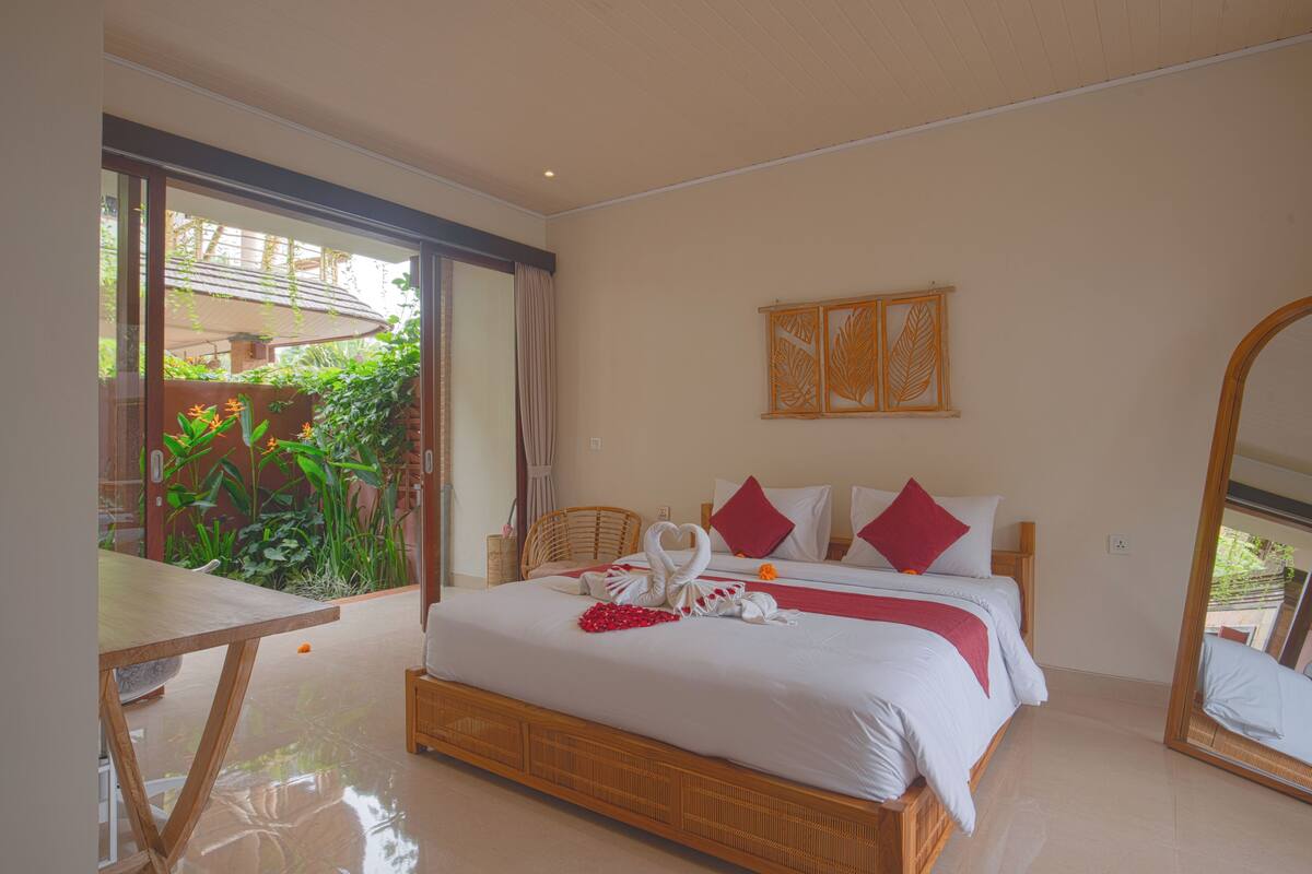 A spacious bedroom with a large bed, a dining table, and a view of greenery through the sliding glass door.