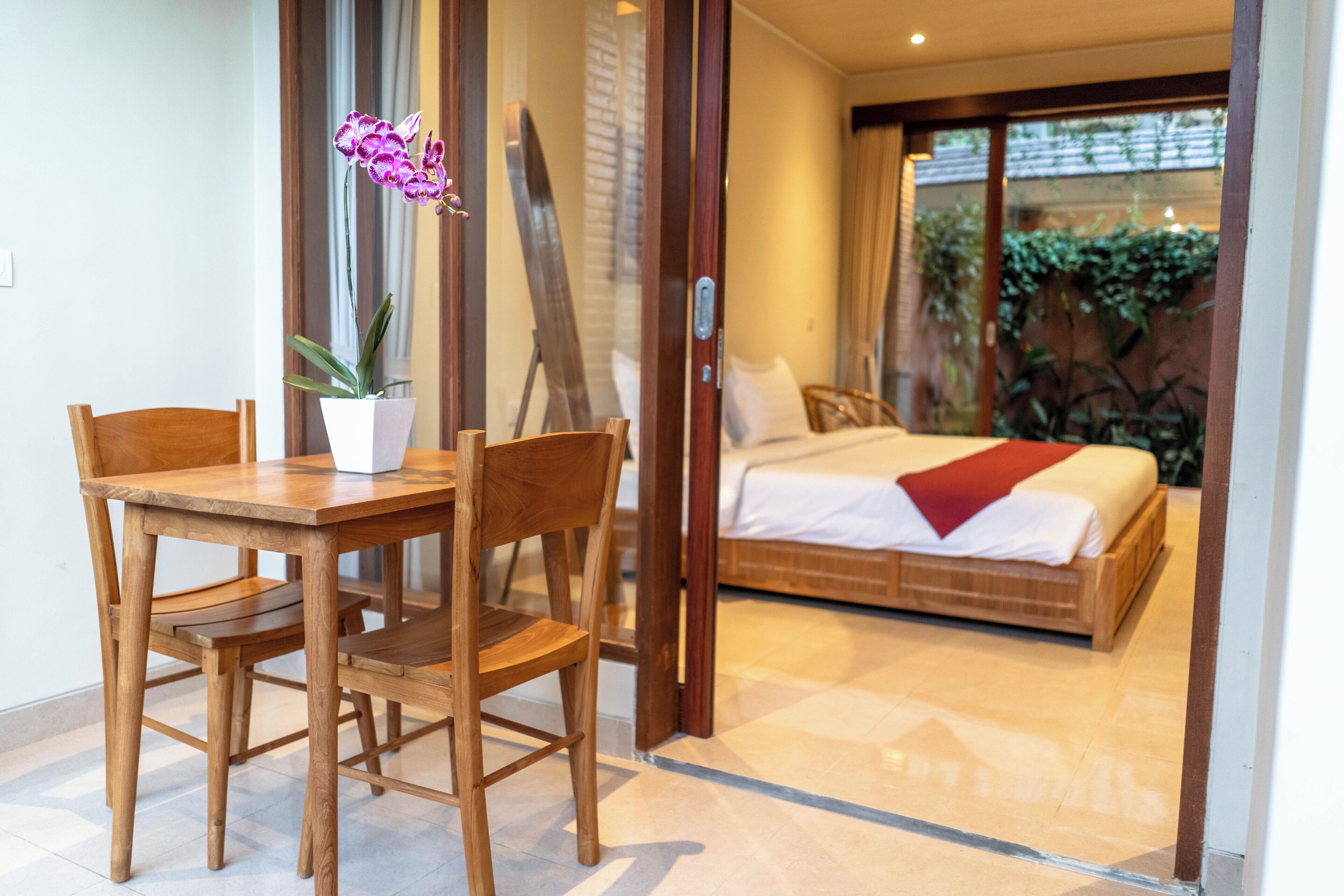 A room with a wooden dining table and chairs, a bed with a red blanket, and a potted orchid.