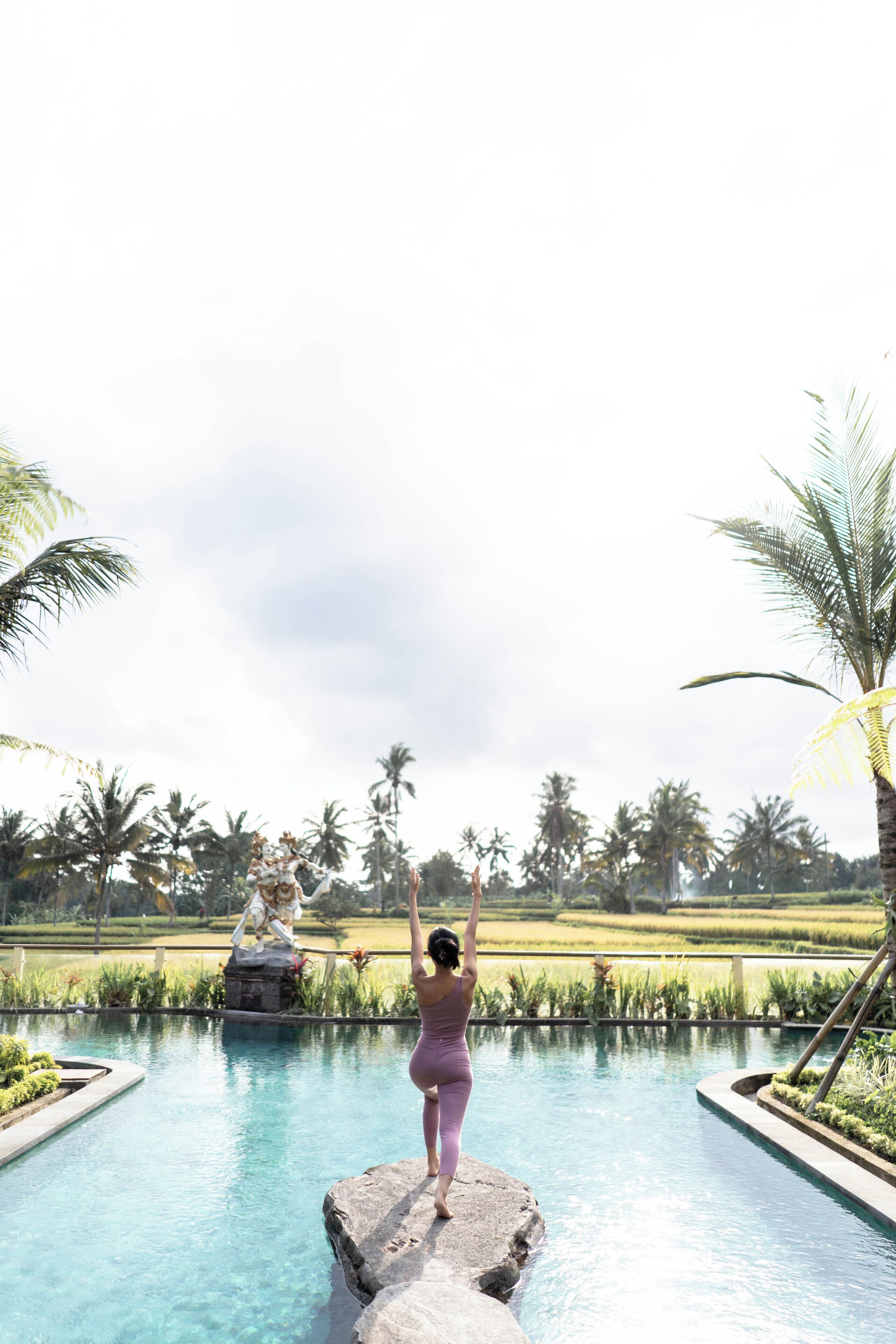 A woman standing on a rock by a pool with palm trees and a statue in the background.