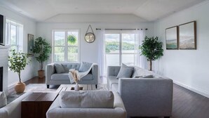 A spacious living room with a fireplace, two grey sofas, a coffee table, and large windows offering a view of the outdoors. - Luxury family retreat on lakefront with breathtaking view, game room, gym &more! (Kawartha Lakes)