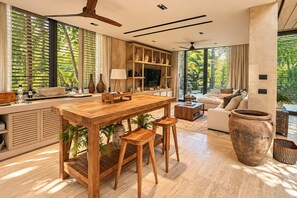 A modern living space with a wooden dining area, a sofa, and a large window with a view of greenery. - Corasol Villa · 4 King Suites & Private Pool (Playa del Carmen)