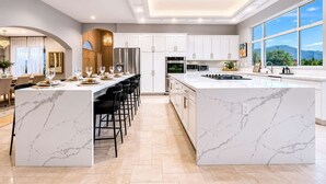 Una cocina moderna con encimeras de mármol, una isla central con taburetes de bar y vistas al exterior a través de amplias ventanas.