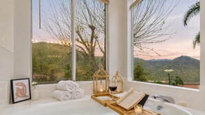 Un baño con bañera, ventana con vistas a árboles y montañas, un cuadro enmarcado y una vela.