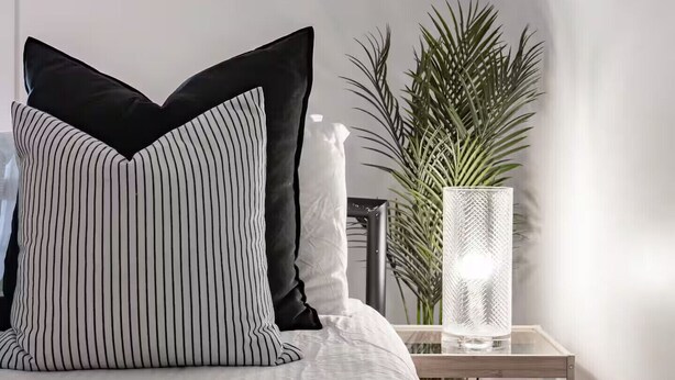 Un lit bien fait, agrémenté d’oreillers à rayures, une petite table de chevet avec une lampe et une plante en pot.