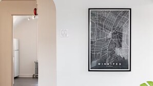 Un intérieur moderne avec une carte encadrée de Winnipeg, un plancher en bois, un mur blanc et une plante.