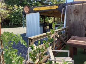 Een houten constructie met een bord waarop de richting naar Coromandel en Tapu staat aangegeven, een rustieke buitenruimte met een houten tafel en stoelen, omringd door groen.