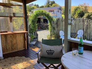 Een houten terras met een groene fauteuil, een ronde tafel met een fles en een glas, en uitzicht op een huis door een houten boog.