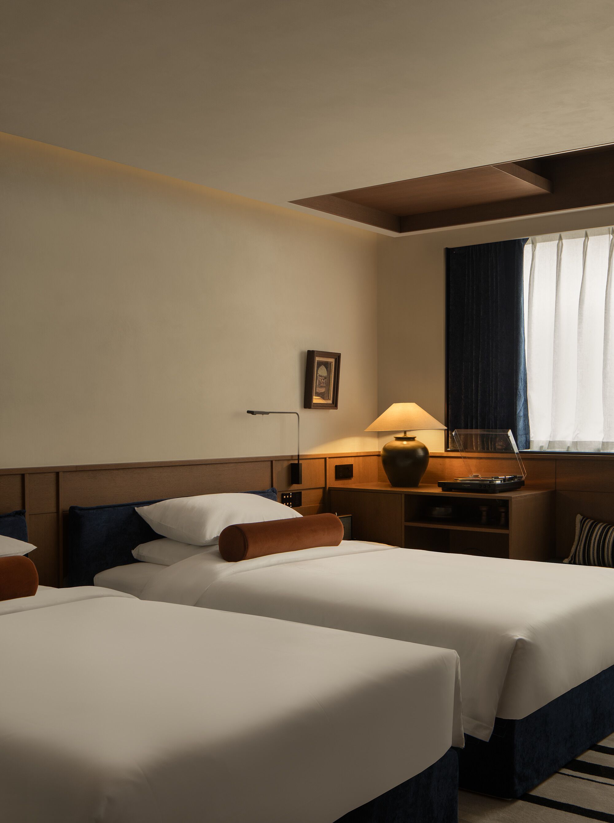 Hotelkamer met twee bedden, een bureau met een lamp, een tafeltje met een vaas en een raam met gordijnen.