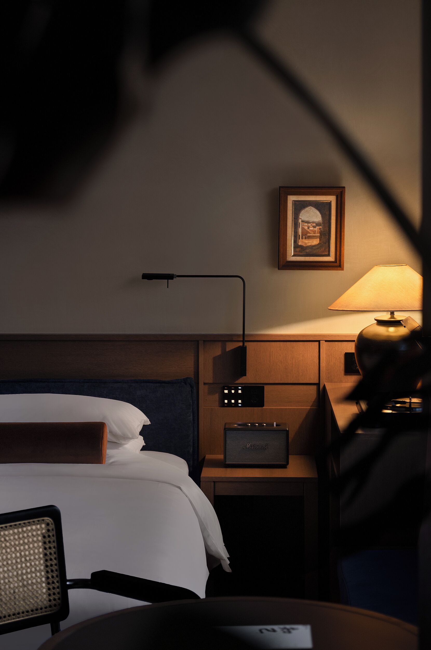 A hotel room with a bed, a desk with a lamp, a chair, a bedside table, and a framed picture on the wall.