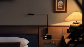 A hotel room with a bed, a desk with a lamp, a chair, a bedside table, and a framed picture on the wall.