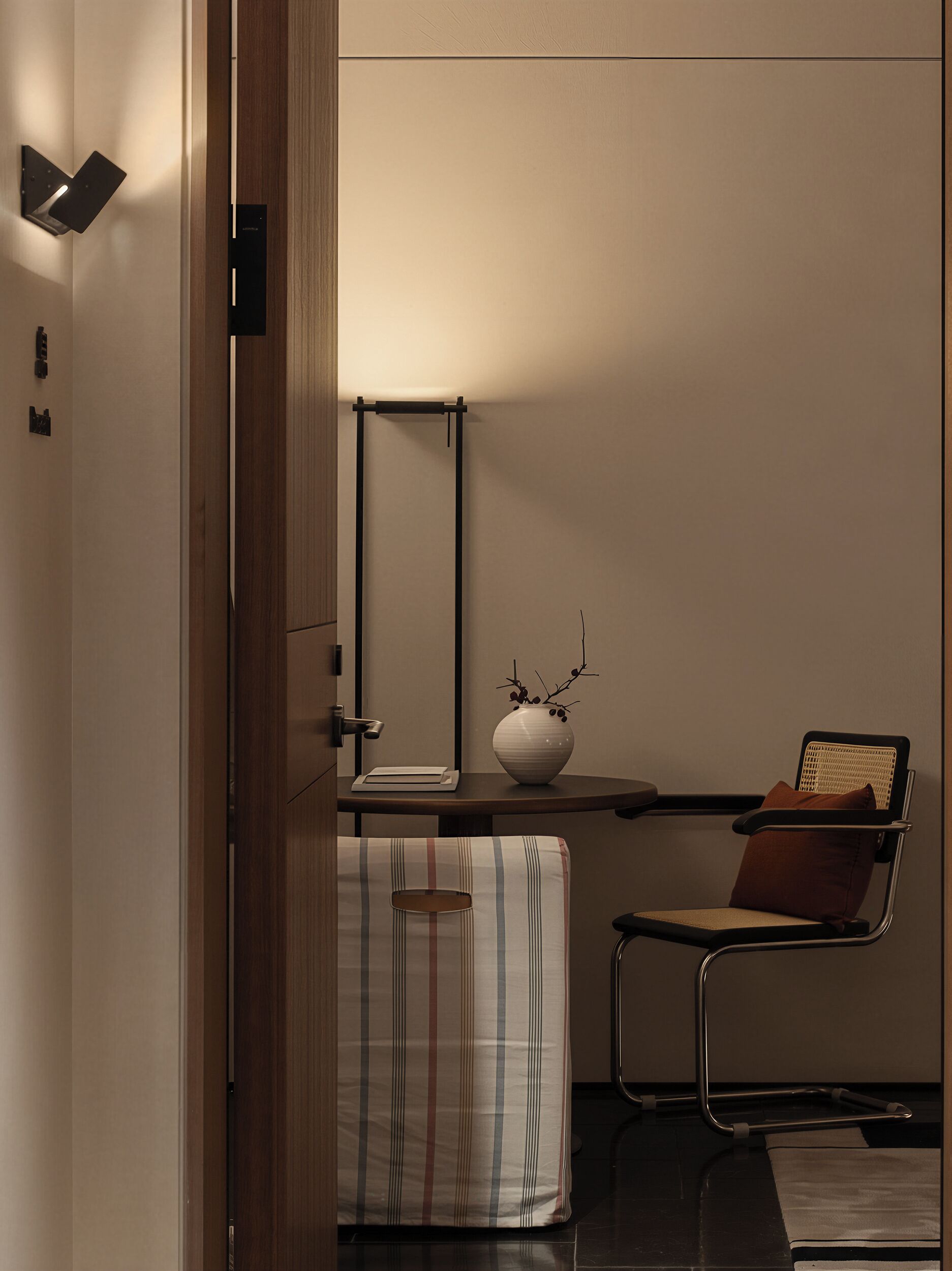 Una habitación moderna con una puerta de madera, un escritorio con lámpara, una silla y un florero con ramas.