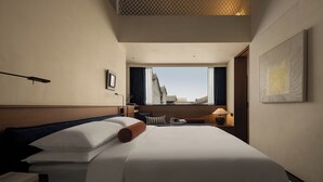 A hotel room with a bed, a desk, a lamp, a window with a view of buildings, and a painting on the wall. - AnHeANAHATA (Datong)
