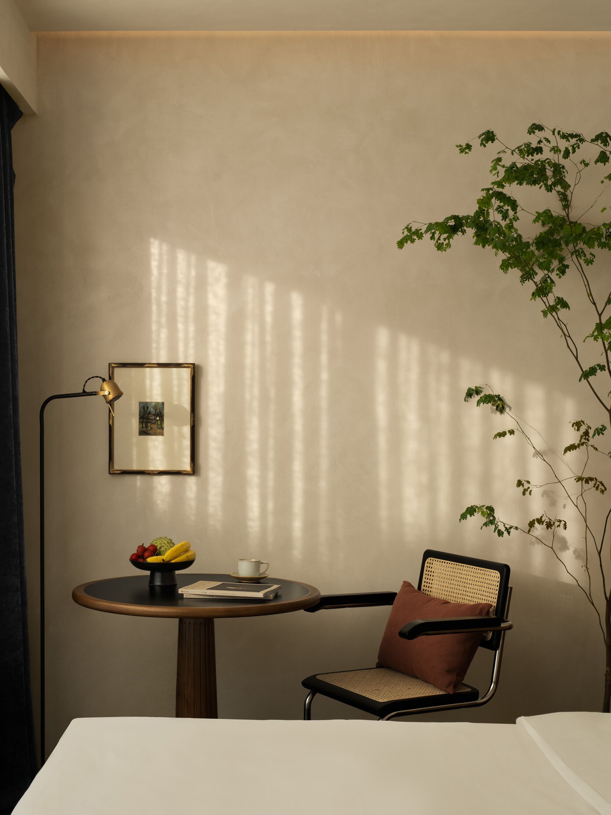 Une chambre d’hôtel avec une table ronde, une chaise, une lampe, un tableau encadré et une plante.