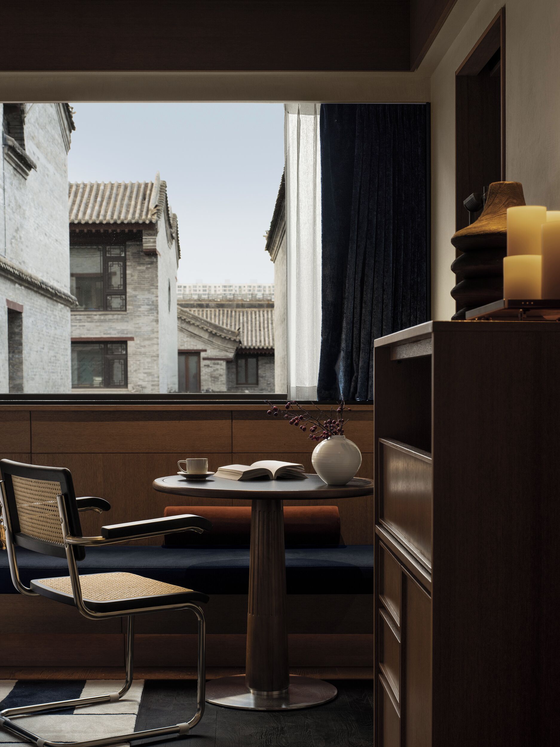 A modern room with a view of traditional buildings, a round table with a book and a vase, a chair, and a wooden cabinet with candles.