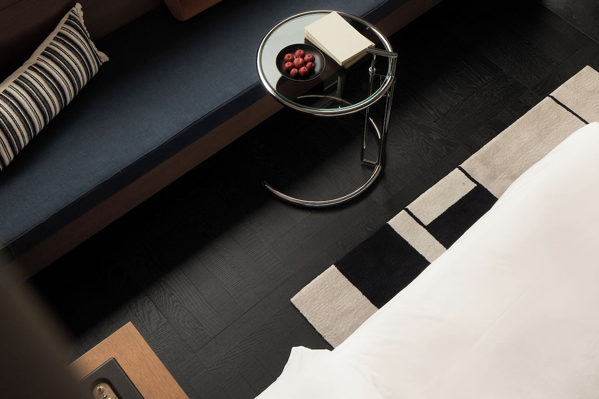 A modern hotel room with a sofa, a small round table with a bowl of fruit, a lamp, and a bed.
