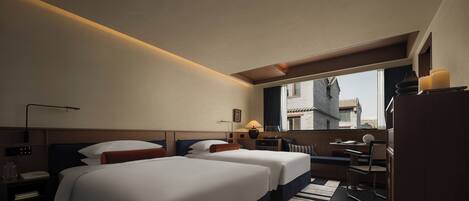 A hotel room with two beds, a desk, a chair, a lamp, and a view of buildings outside the window.