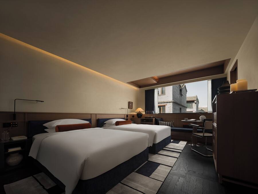 Hotelkamer met twee bedden, een bureau, een stoel, een lamp en uitzicht op gebouwen.