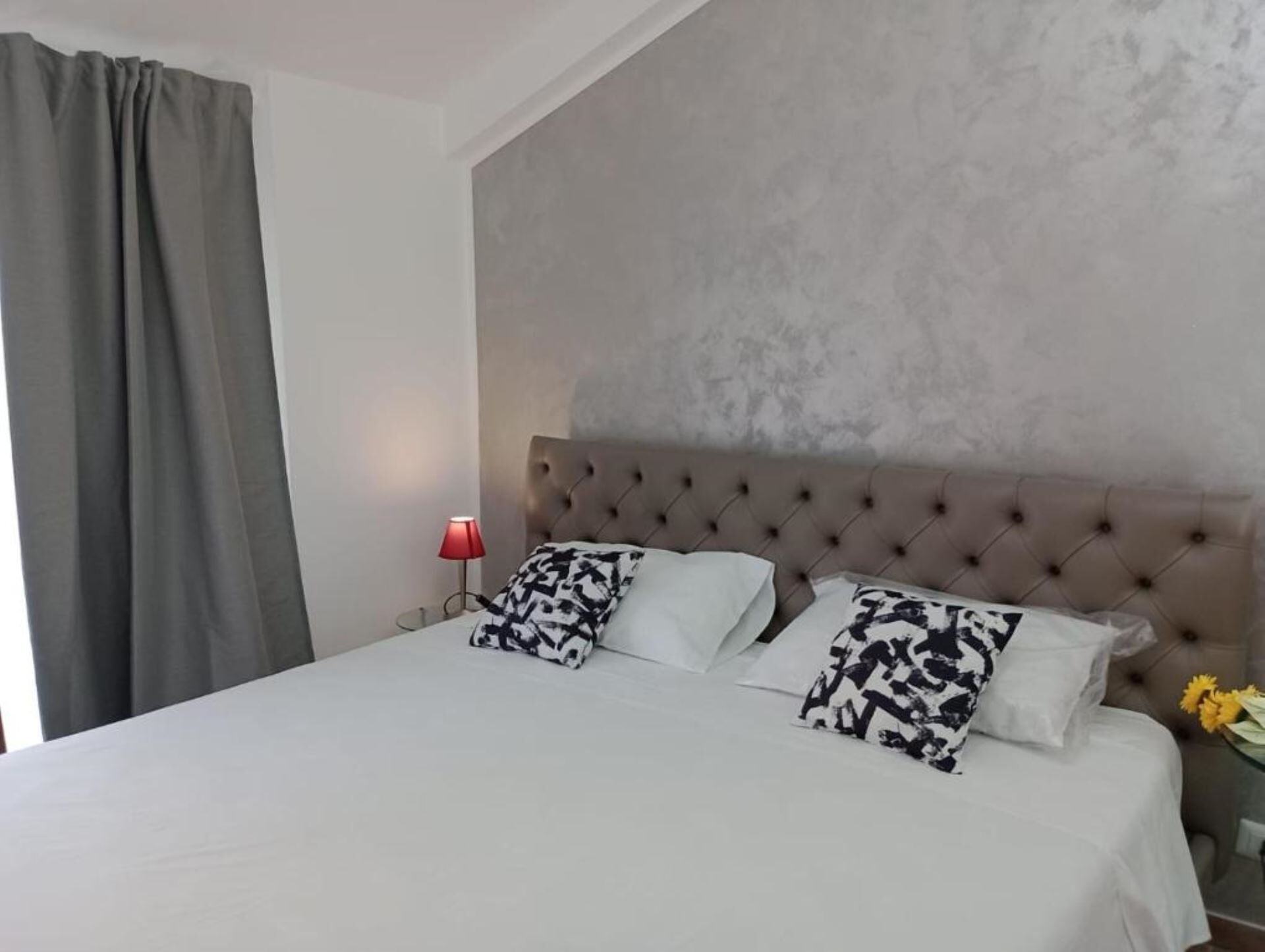 Una cama bien tendida con un cabecero capitonado, dos almohadas estampadas, una lámpara de noche roja y una cortina gris.