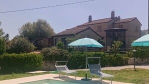 Una piscina con sillones de descanso y sombrillas, una casa de ladrillo al fondo y un cielo despejado.