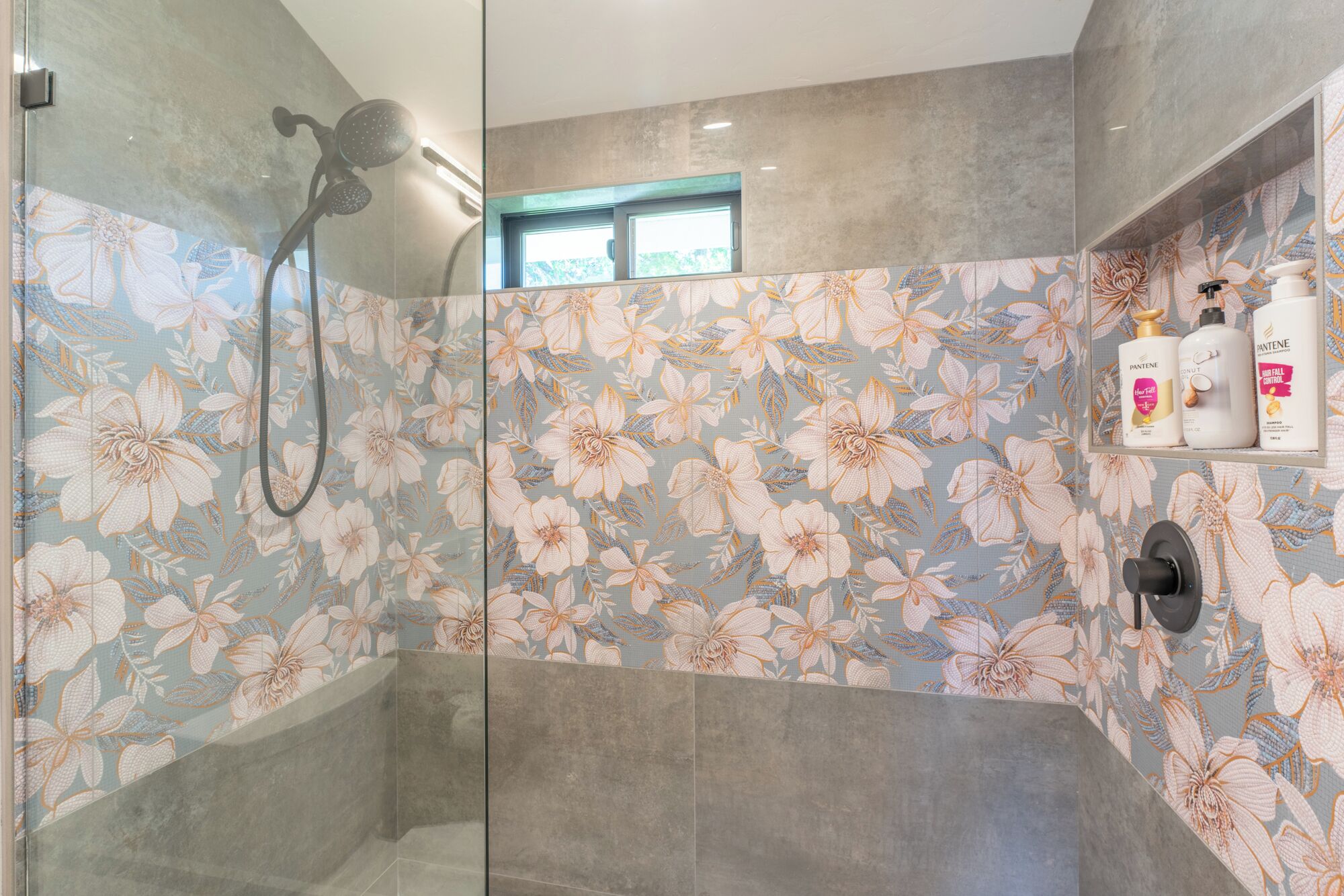 A shower with a floral tile design, a glass door, and a shelf with toiletries.