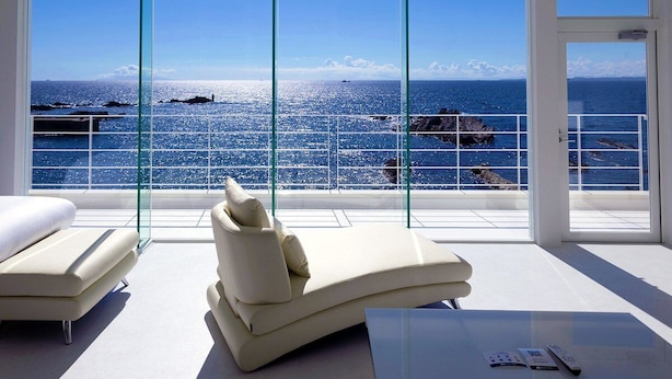 Una habitación moderna con un amplio ventanal con vistas al mar, dos sillones blancos y una mesita con un control remoto.