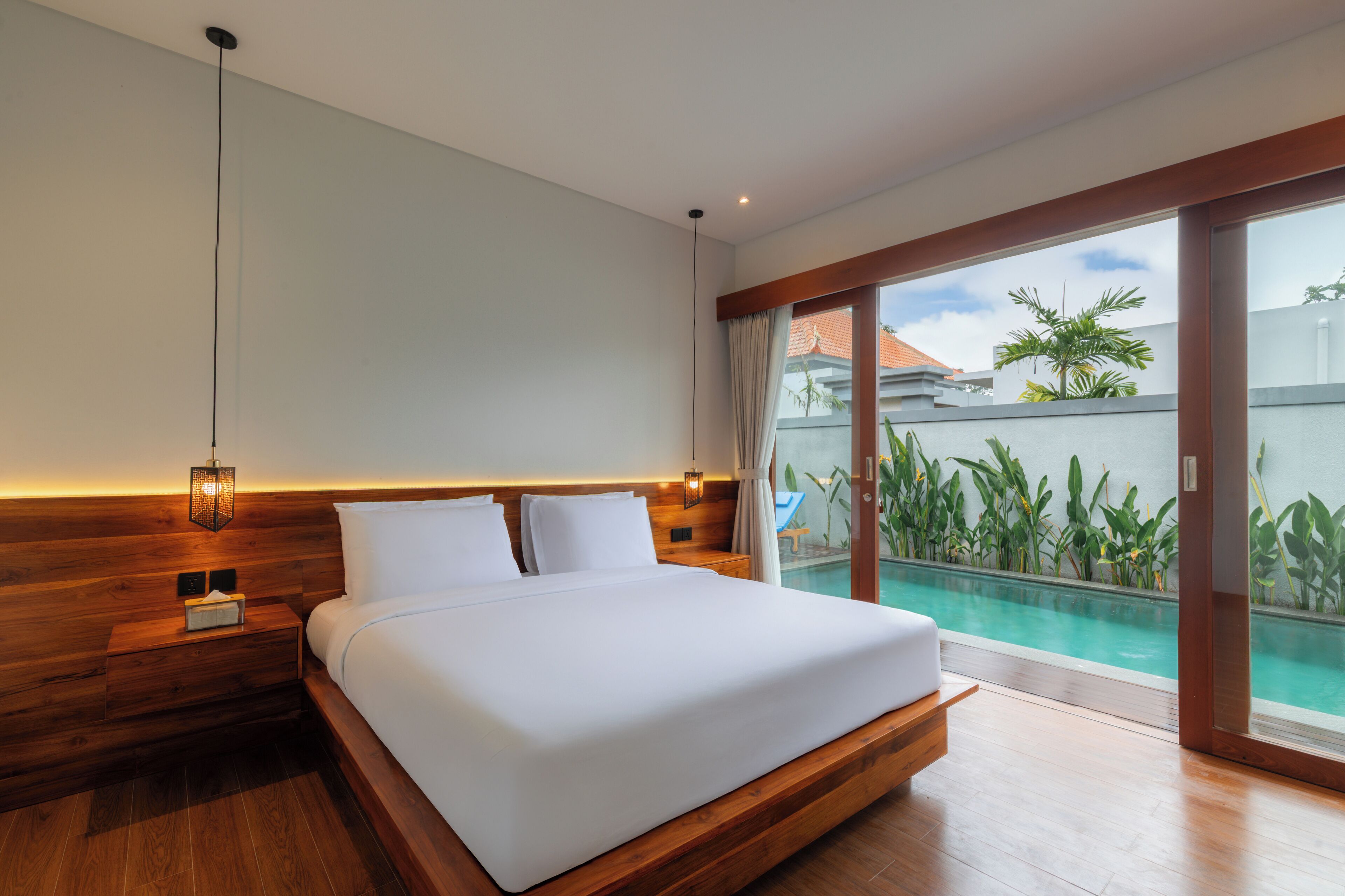 Habitación de hotel moderna con una cama grande, cabecera de madera, una mesita de noche, una lámpara y vista a una piscina a través de una puerta de cristal.