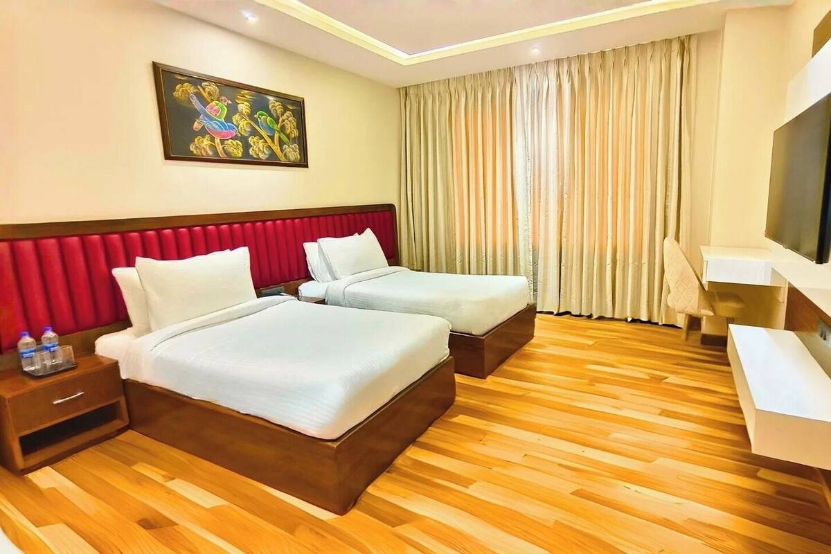 A hotel room with two beds, a red headboard, wooden flooring, a painting on the wall, and a TV on the wall.