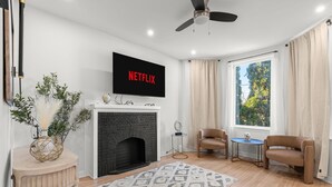 Ein Wohnzimmer mit Kamin, einem darüber montierten Fernseher, zwei Ledersesseln, einem kleinen runden Tisch und einem großen Fenster mit Vorhängen.