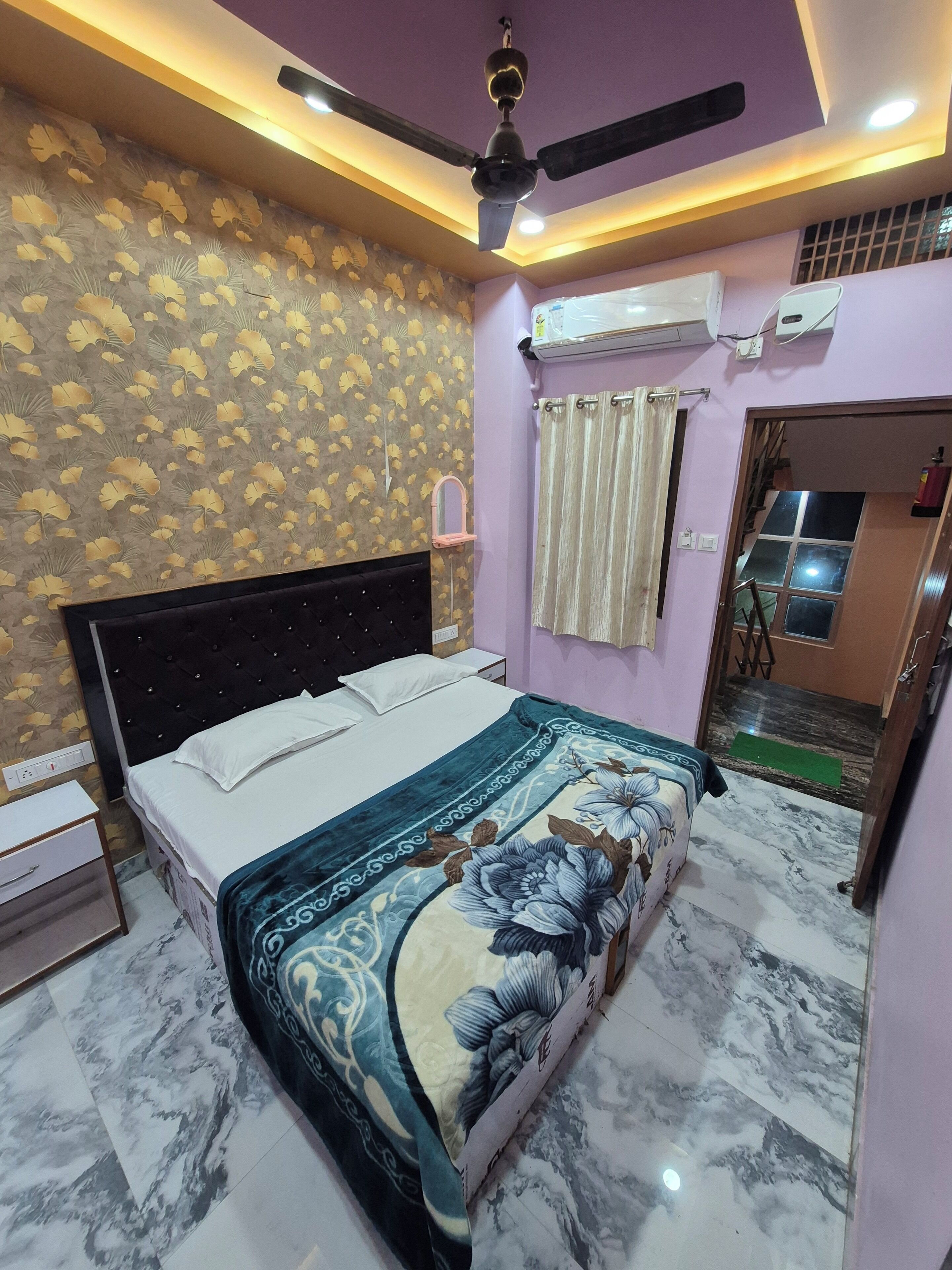 A bedroom with a bed, a ceiling fan, an air conditioning unit, and a bathroom visible through an open door.