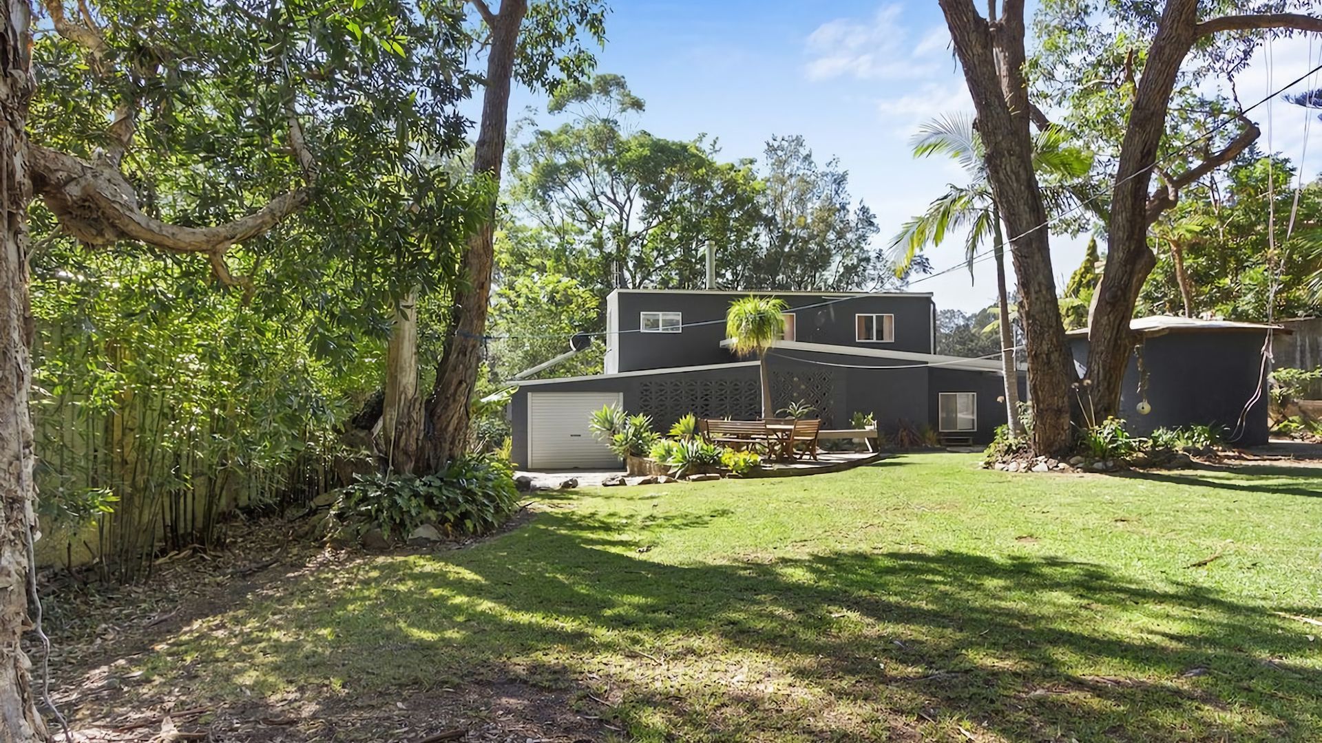A modern two-story house with a covered patio, surrounded by lush greenery and trees.