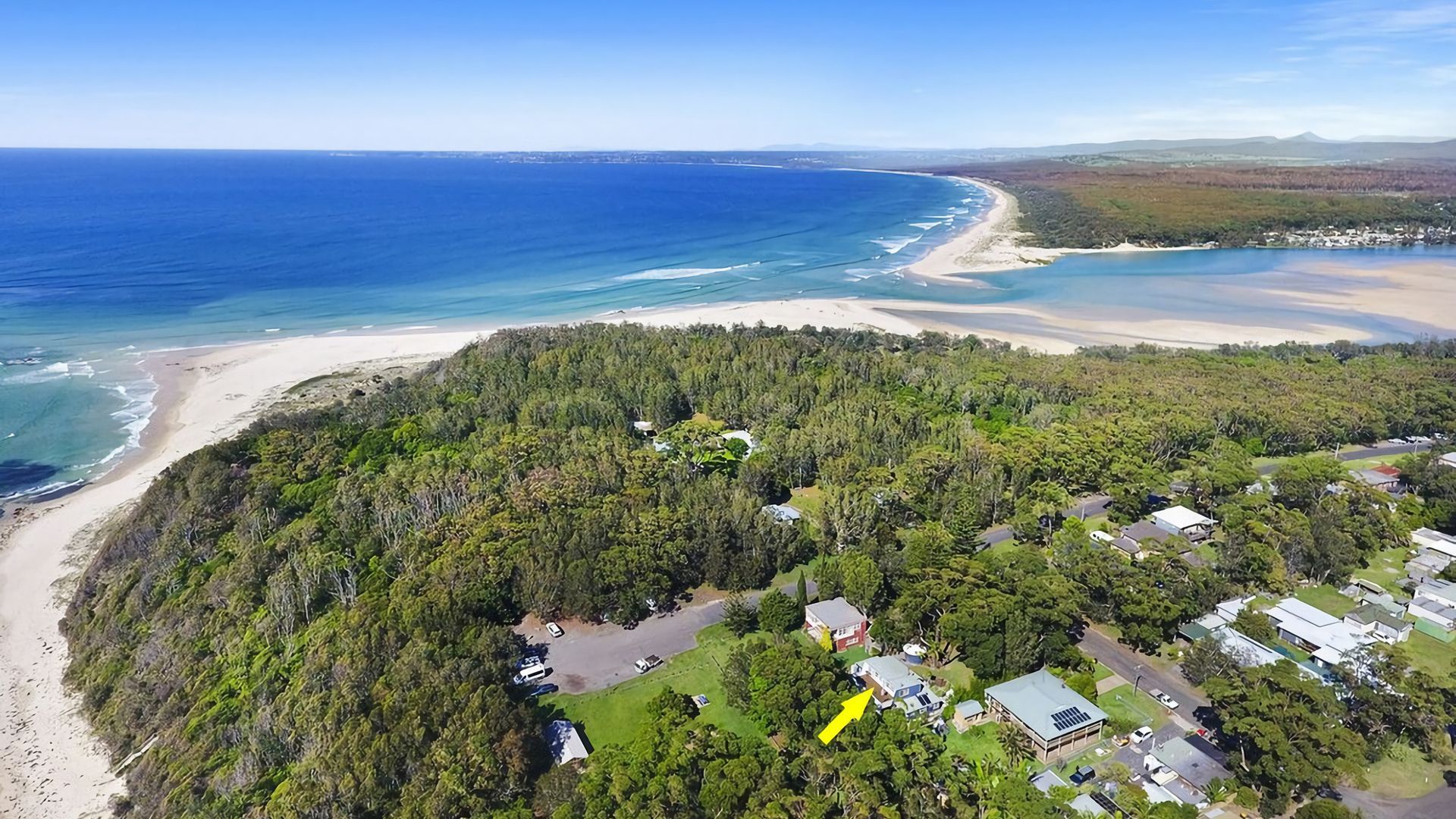 An aerial view of a coastal area with a beach, dense forest, and residential buildings.