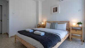 A bedroom with a wooden bed, white bedding, a dark throw blanket, two bedside tables with lamps, a wall-mounted air conditioner, and a door. - Blossom 1-BR Apt. & Balcony in Larnaca (Larnaca)
