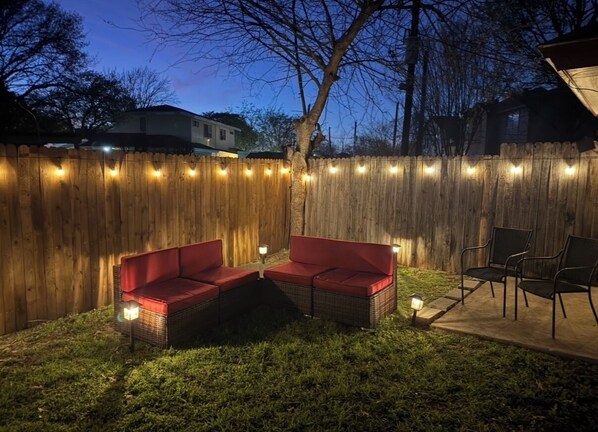 Un patio trasero con valla de madera, dos sillones rojos acolchados, una mesa de mimbre y luces colgantes.