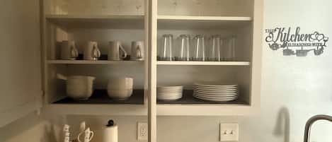 Una cocina con estantes blancos que sostienen vasos, cuencos y platos. Hay un bloque de cuchillos, un dispensador de toallas de papel y un grifo.