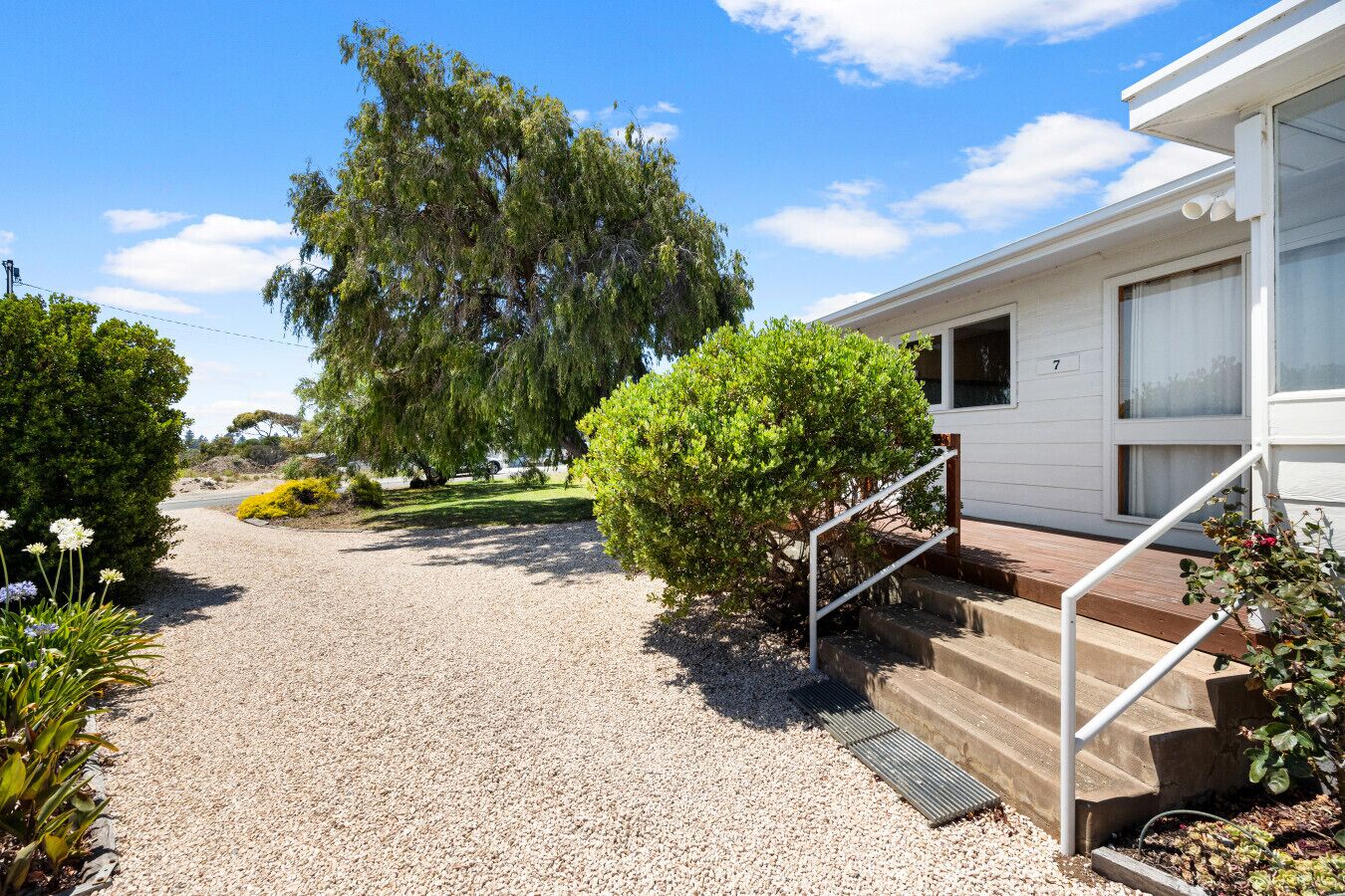 A gravel driveway leading to a white building with a number '7' on the door. There are steps with a metal handrail, a small bush, and a tree in the background.