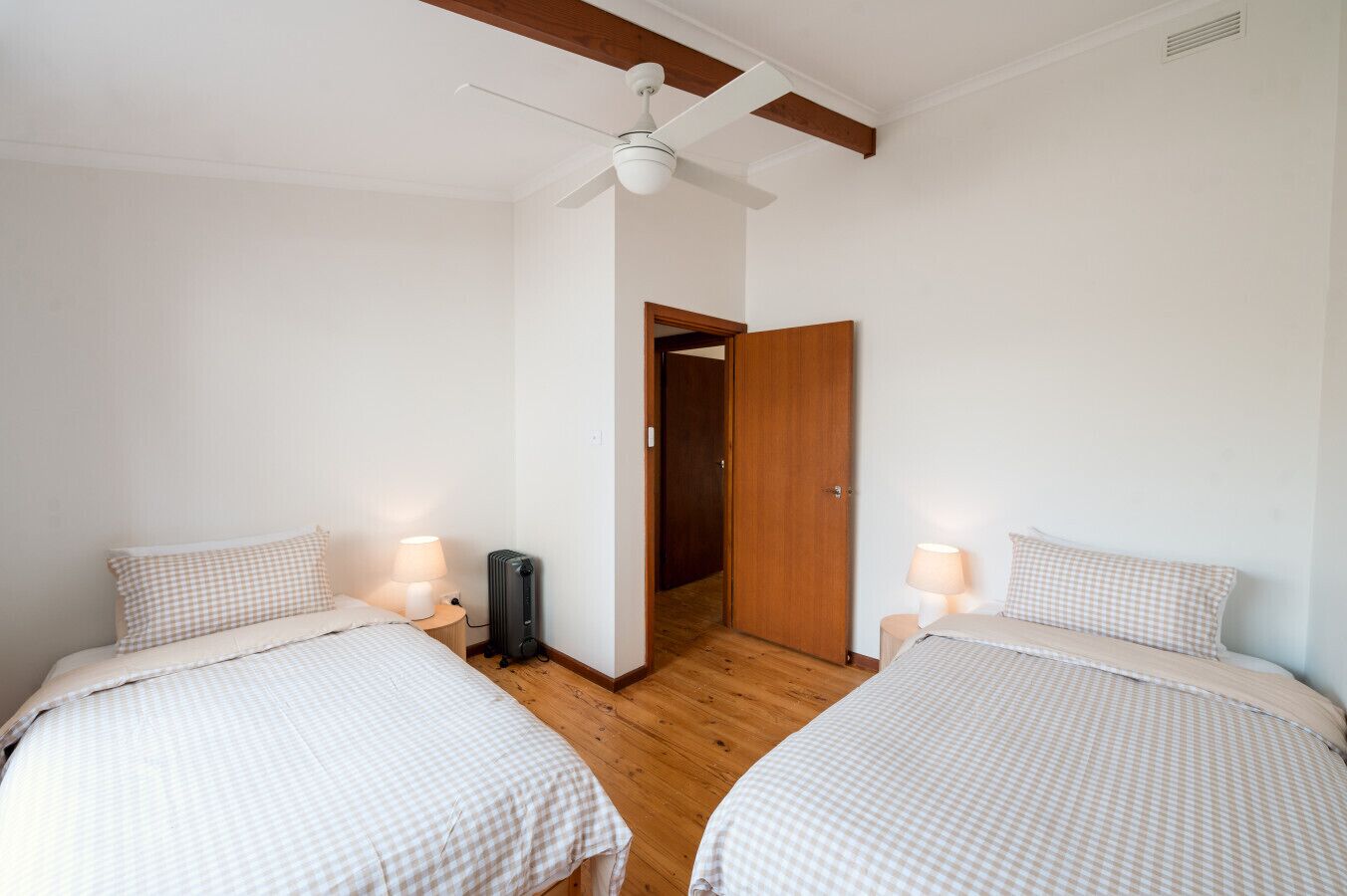 Ein Zimmer mit zwei Betten, jeweils mit karierter Bettdecke und Nachttischlampe. Es gibt einen Deckenventilator, eine Heizung und eine Holztür zu einem anderen Raum.
