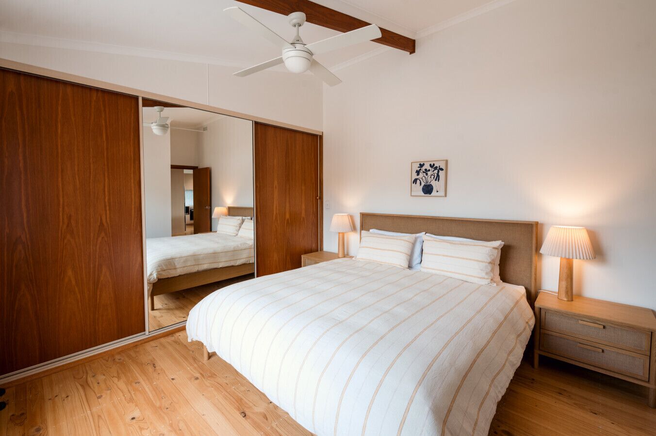 A bedroom with a large bed, wooden flooring, a ceiling fan, a bedside table with a lamp, and a mirror.