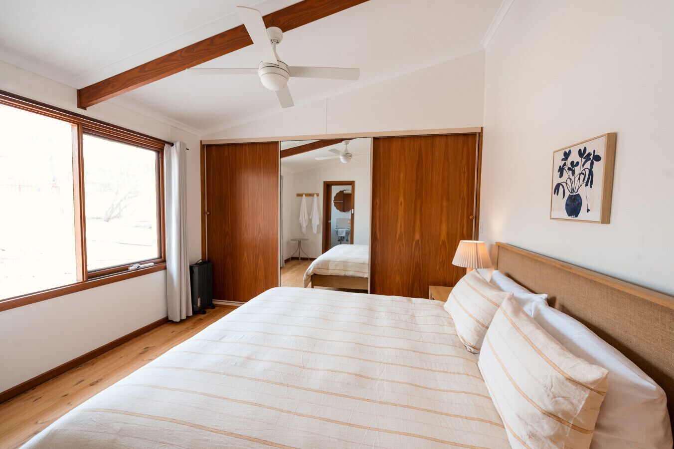 A bedroom with a large bed, wooden ceiling beams, a ceiling fan, a window, and a mirror.