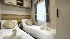 Quarto de hotel com duas camas, uma cabeceira de madeira, uma mesinha de cabeceira com uma planta e uma janela com cortinas.