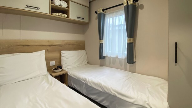 Une chambre avec deux lits simples, une tête de lit en bois, une petite table de chevet avec un bol, et une fenêtre avec des rideaux.