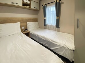 Een kamer met twee eenpersoonsbedden, een houten hoofdbord, een klein nachtkastje met een schaal, en een raam met gordijnen.